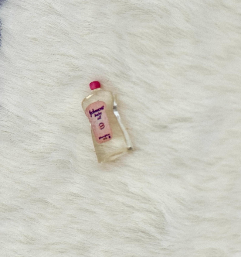 Może przedstawiać: Miniaturowa butelka z przezroczystego plastiku z oliwką dla dzieci Johnson's, posiadająca r&oacute;żową etykietę z napisem Johnson's baby oil wykonanym ciemną czcionką oraz małą r&oacute;żową nakrętkę. Butelka ma zakrzywiony kształt i jest wykonana jako replika.