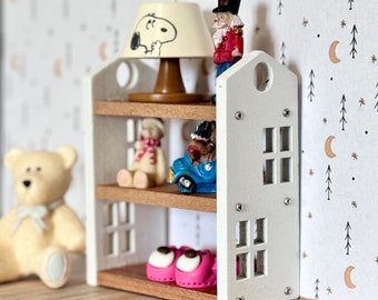 May include: A white wooden shelf unit with three shelves, featuring a variety of miniature toys, including a teddy bear, a toy car, a nutcracker, and a lamp with a Snoopy-themed lampshade. The shelf unit is against a white background with a repeating pattern of brown trees and crescent moons.