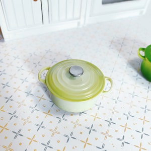 May include: A small green enamelled cast iron pot with a silver knob on the lid. The pot is sitting on a white and grey tiled surface.
