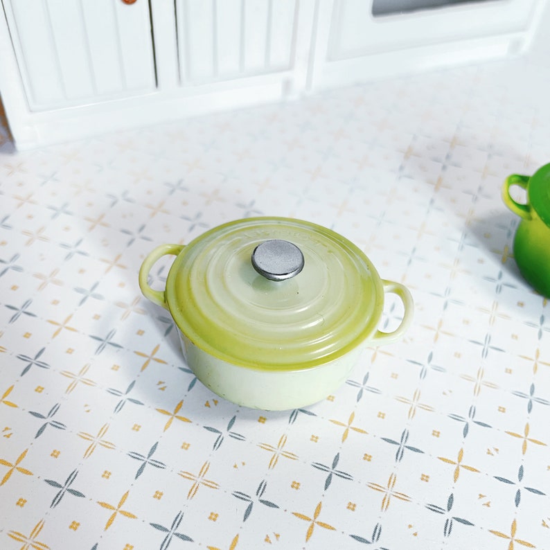 May include: A small green enamelled cast iron pot with a silver knob on the lid. The pot is sitting on a white and grey tiled surface.