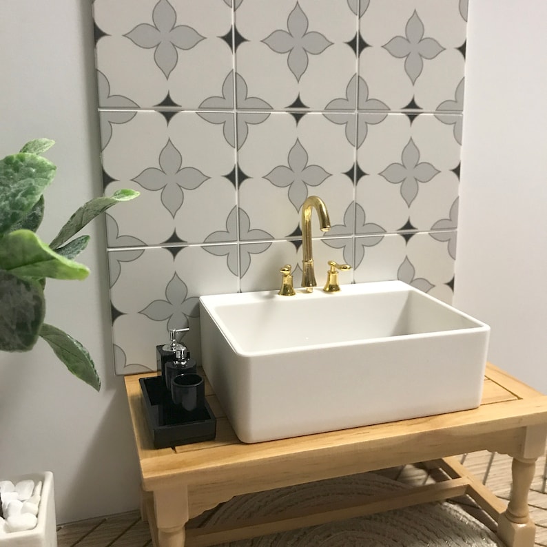 May include: A white porcelain sink with a gold faucet and two handles sits on a wooden countertop with a black tray holding two bottles and a glass. The sink is in front of a white tiled wall with a gray and black geometric pattern.