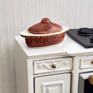May include: Miniature brown and white ceramic casserole dish with a lid. The oval-shaped dish has a decorative pattern around the rim. The lid has a small handle. The dish is on a white cabinet, suggesting a kitchen or dining theme.