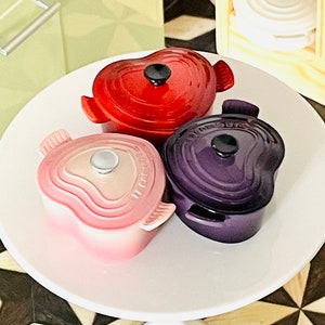May include: Three heart-shaped mini Dutch ovens in red, pink, and purple. The ovens are sitting on a white pedestal table.