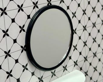 May include: A round black-framed mirror hangs on a wall with a black and white geometric tile pattern. A white shelf is positioned below the mirror. The mirror reflects the room.