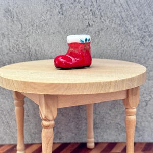 May include: A miniature red Christmas stocking with a white cuff and blue and green accents. The small ceramic stocking is displayed on a small, round wooden table with four legs. The table is made of light-coloured wood.