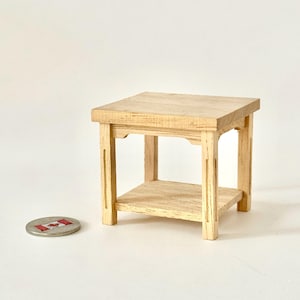May include: A miniature wooden side table with a square top and a lower shelf. The table is made of light-coloured wood and has a simple, classic design. The table is approximately 7.6 cm tall.