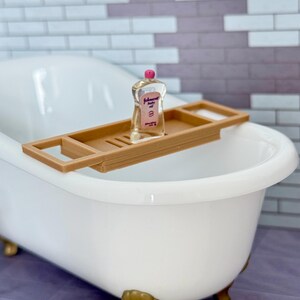 Może przedstawiać: Miniaturowa biała wanna na złotych n&oacute;żkach z brązową, prostokątną p&oacute;łką łazienkową umieszczoną na g&oacute;rze. Na środku p&oacute;łki znajduje się mała, przezroczysta plastikowa butelka oliwki dla dzieci Johnson's z r&oacute;żową nakrętką.