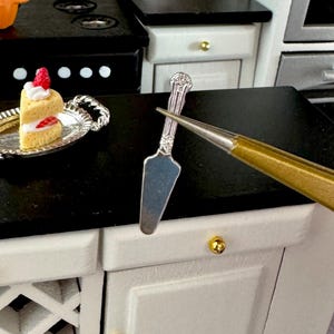 May include: Miniature kitchen scene with a slice of cake on a silver plate, a silver cake server, and a small mug with a fall-themed design. The kitchen features a black countertop and white cabinets with gold knobs.
