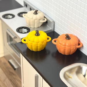 Puede incluir: Tres macetas de cerámica en miniatura con forma de calabaza en blanco, amarillo y naranja. Las macetas están sobre una encimera negra en una cocina de casa de muñecas.