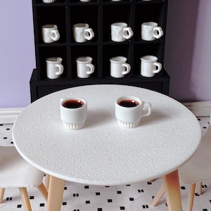 Può includere: Tazzine da caffè in ceramica bianca in miniatura, alcune riempite con liquido scuro, sono disposte su un tavolo bianco e in un mobile nero. Il tavolo ha gambe in legno chiaro. La scena suggerisce una casa delle bambole o un'ambientazione in miniatura.