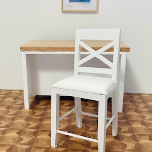 May include: A miniature white wooden chair with an X-shaped back and a white cushioned seat. The chair is positioned in front of a small desk with a light wood top and white legs. A framed artwork hangs on the wall.