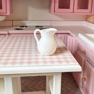 Puede incluir: Una jarra de cerámica blanca con un diseño de espiral se encuentra sobre una encimera de gingham rosa y blanco en una cocina de casa de muñecas.