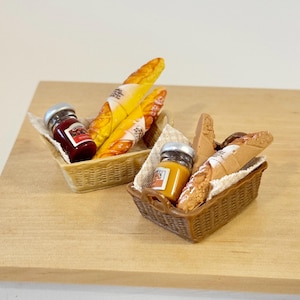 Può includere: Due cestini in miniatura in vimini pieni di generi alimentari. Un cestino contiene un vasetto di marmellata rossa e pane avvolto. L'altro cestino ha un vasetto di marmellata gialla e pane. I cestini sono su una superficie marrone chiaro.