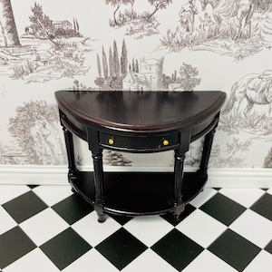 Puede incluir: Una mesa negra de media luna con un cajón y un estante inferior. La mesa tiene patas torneadas y herrajes de color dorado. La mesa está sobre un suelo de baldosas blancas y negras.