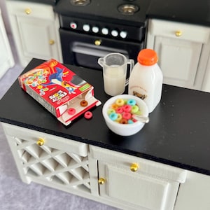 Puede incluir: Escena de cocina en miniatura con encimera negra. Cuenta con una caja de cereales Froot Loops, un cuenco de cereales, una jarra de leche y una botella de leche. La cocina tiene armarios blancos con pomos dorados y una estufa negra.