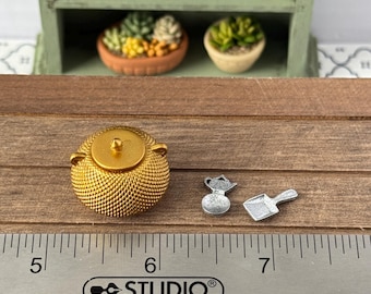 May include: A gold-colored miniature teapot with a textured surface, a silver-colored miniature watering can, and a silver-colored miniature dustpan. The items are on a wooden surface with a ruler marked in inches.