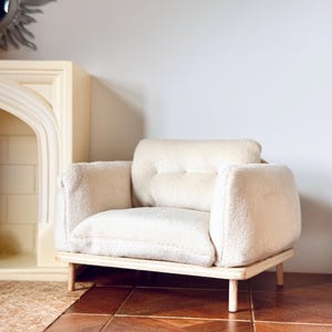 May include: A white plush armchair with a wooden base and four legs. The chair is sitting on a wooden floor with a rug in the foreground.