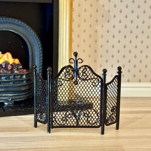 Peut inclure: Un écran de cheminée miniature en métal noir doté d'un design pliable à trois panneaux avec un motif de grille en maille fine, des volutes décoratives en haut et en bas, et quatre montants verticaux surmontés de petits fleurons sphériques.