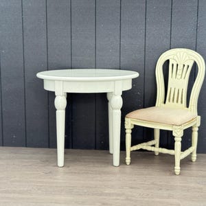 Op de afbeelding: Een kleine, witte, ronde tafel met gedraaide poten staat naast een crèmekleurige stoel met een patroon op de rug en een roze zitkussen. De meubels staan tegen een donkergrijze wand met panelen en een lichte houten vloer.