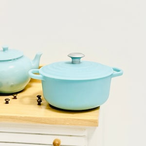 May include: A light blue enamel cast iron pot with a silver knob on the lid. The pot is sitting on a wooden surface.