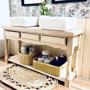 以下が含まれることがあります： 2 つのシンクと引き出し付きの木製ベースが付いた白い洗面台。洗面台は、織りの敷物、タオルのバスケット、トイレタリーのバスケットで飾られています。