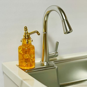 May include: A miniature kitchen scene featuring a silver faucet and a clear, yellow soap dispenser with a gold pump. The dispenser is filled with a yellow liquid and sits next to a stainless steel sink.