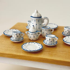 Può includere: Servizio da tè in miniatura in porcellana bianca con pois blu e bordi blu. Il set comprende una teiera, una lattiera, una zuccheriera, tazze, piattini e piatti. Il servizio da tè è esposto su una superficie di legno.