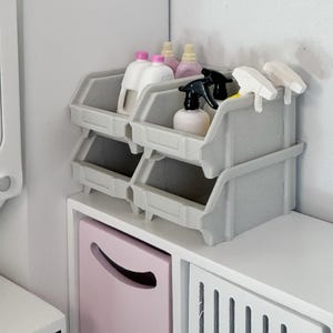 Puede incluir: Cuatro contenedores de almacenamiento de plástico gris apilados uno encima del otro. Los contenedores están llenos de varias botellas de productos de limpieza, incluyendo una botella rosa, una botella amarilla y una botella blanca. Las botellas tienen pulverizadores negros y blancos.