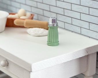 May include: A miniature green salt shaker with a silver top sits on a white table. The shaker has a ribbed design and is part of a kitchen-themed display. Other items include a rolling pin, dough, and a basket of eggs.