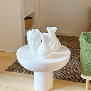 May include: Three white ceramic vases of different shapes and sizes are arranged on a white pedestal table. The table has a textured surface and a round top. The vases are all simple and modern in design.