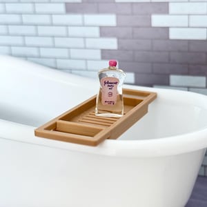Może przedstawiać: Miniaturowa, przezroczysta plastikowa butelka oliwki dla dzieci Johnson's z r&oacute;żową nakrętką i etykietą, umieszczona na małej, brązowej, drewnianej p&oacute;łce łazienkowej z listewkami, położonej na krawędziach białej miniaturowej wanny.