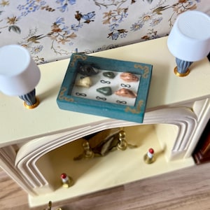 Peut inclure: Une vitrine miniature turquoise contenant diverses petites pierres colorées est posée sur une cheminée crème. Deux petites lampes blanches avec des bases bleues et dorées encadrent l'exposition. La cheminée a une ouverture décorative.