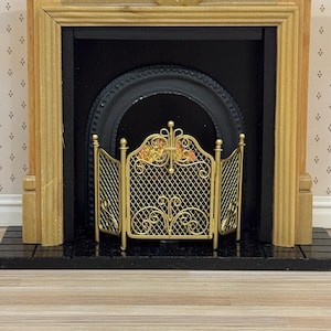 Peut inclure: Une cheminée miniature avec un manteau en bois clair, un foyer arqué noir et un écran pliant à trois panneaux en métal doré avec un motif en maille losange et des volutes décoratives, posée sur une base de foyer noire.