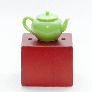 May include: A small green teapot with a lid sits on a red wooden block.