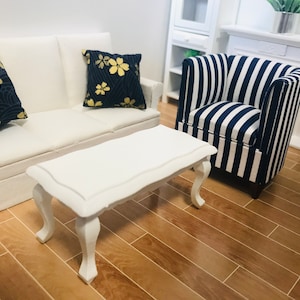 May include: Miniature living room set featuring a white sofa, a white coffee table, and a striped armchair. The sofa has two navy blue cushions with gold floral designs. The coffee table has curved legs.