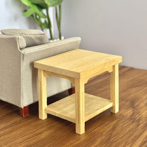 May include: A miniature, light-coloured wooden side table with a lower shelf. The table is square, with a simple design and visible wood grain. It is placed next to a beige sofa. The table measures approximately 15 cm in length and width.