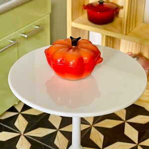 May include: A red pumpkin-shaped ceramic casserole dish with a black knob on the lid. The dish is sitting on a white table.
