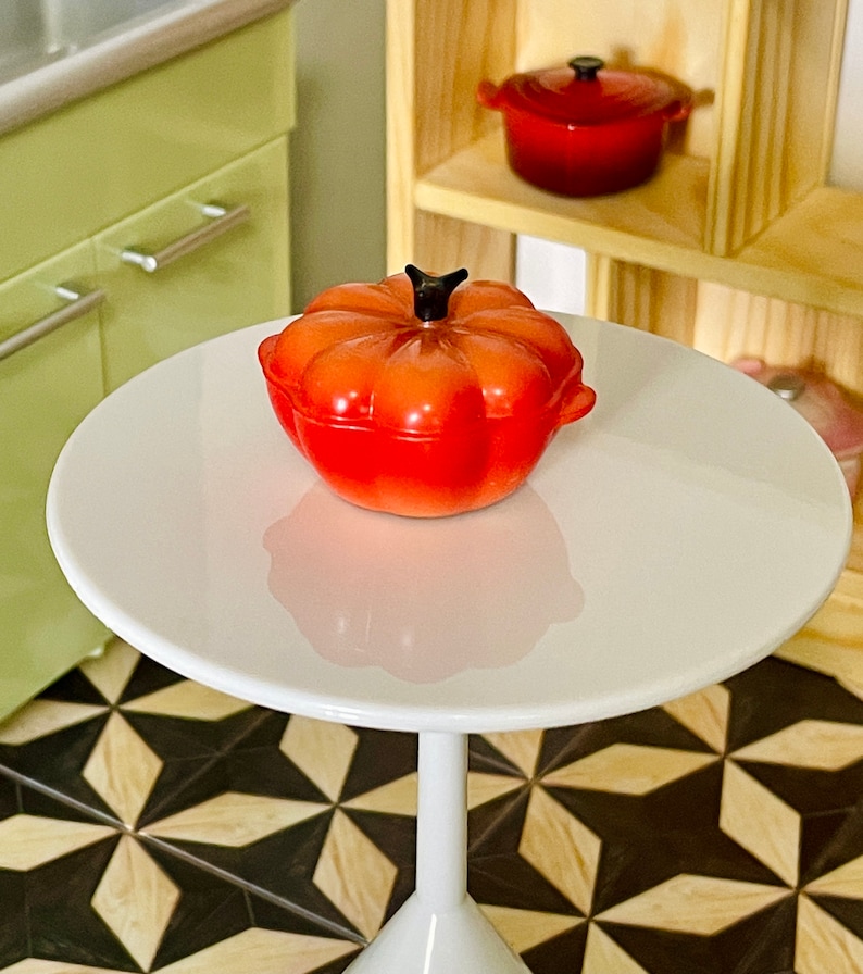 May include: A red pumpkin-shaped ceramic casserole dish with a black knob on the lid. The dish is sitting on a white table.