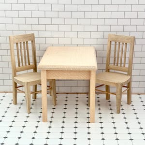 May include: A miniature wooden dining set comprising a small square table and two matching chairs. The furniture is light-coloured, with a natural wood grain finish. The chairs have vertical back slats.
