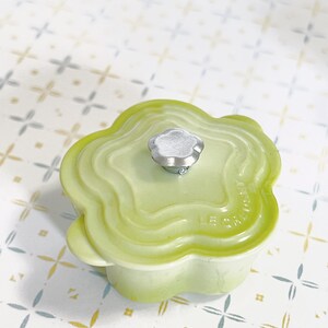 May include: A light green ceramic pot with a flower-shaped lid and a silver knob. The pot has a raised design around the rim and the words "Le Creuset" are visible on the side.