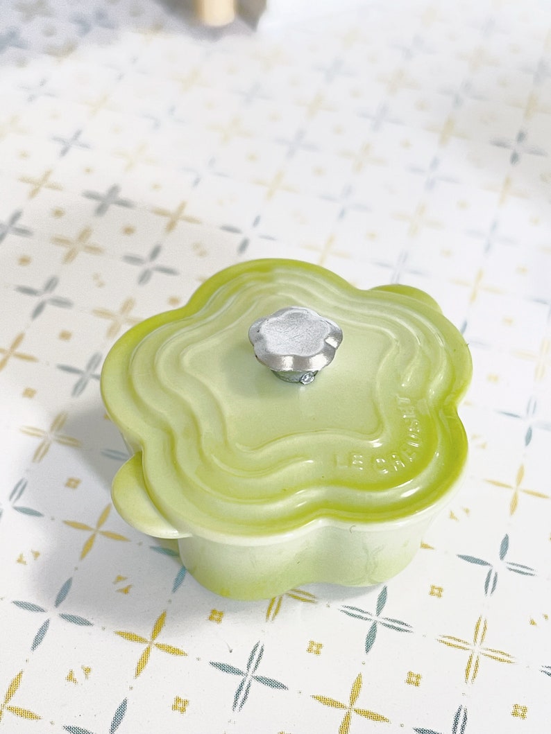May include: A light green ceramic pot with a flower-shaped lid and a silver knob. The pot has a raised design around the rim and the words "Le Creuset" are visible on the side.