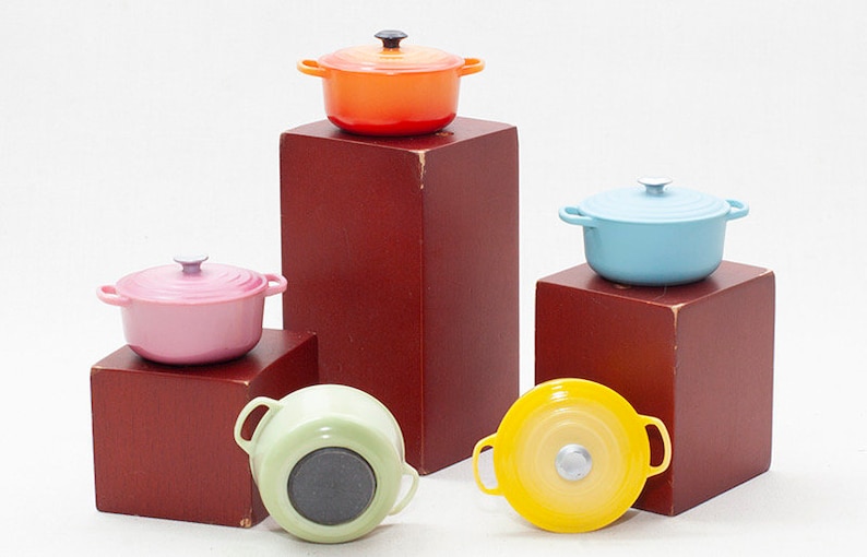 May include: Five miniature cast iron pots in different colours: orange, blue, pink, green and yellow. The pots are arranged on wooden blocks.