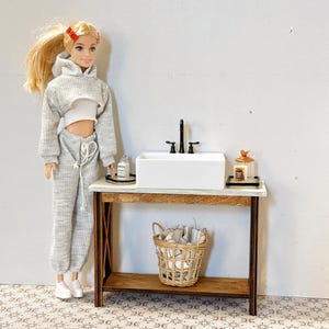 May include: A dollhouse bathroom vanity set with a white porcelain sink, black faucet, and a wooden countertop. The vanity has a lower shelf with a wicker basket and a small jar on the top shelf. The vanity is in front of a white wall with a tiled floor.