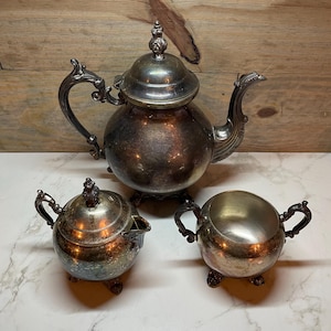 May include: A vintage silver tea set, including a teapot with a lid, a sugar bowl with a lid, and a creamer. The set has ornate handles and rests on small feet. The silver has a tarnished appearance, giving it a classic, antique look.
