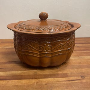 May include: A glossy, brown ceramic casserole dish with a matching lid. The exterior features a raised, embossed scrollwork pattern around the middle and a scalloped rim. The lid has a central knob handle and a matching embossed decorative border.