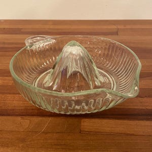 May include: A clear glass citrus juicer with a ribbed bowl and a central reamer. The juicer has a handle and is designed for easy citrus fruit juicing. The glass is transparent, allowing visibility of the fruit and juice.