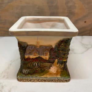 May include: A square ceramic planter with a cottage scene. The planter has a cream-colored rim and a detailed landscape design featuring a thatched-roof cottage, trees, and a stone wall. The base is a stone-like texture.