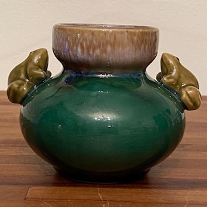 May include: A ceramic vase with a green base and a brown rim. Two frog figurines are attached to the sides of the vase. The vase has a glossy finish and is sitting on a wooden surface.