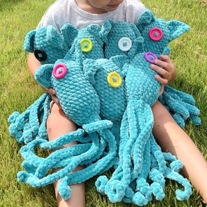 Puede incluir: Una colecci&oacute;n de pulpos de ganchillo turquesa con ojos de bot&oacute;n de varios colores. Los peluches tienen largos tent&aacute;culos rizados y los sostiene una persona sentada sobre hierba verde.