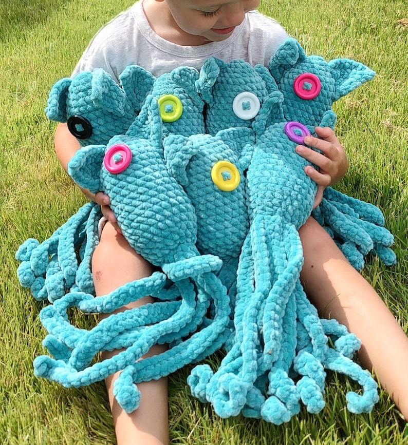 Puede incluir: Una colecci&oacute;n de pulpos de ganchillo turquesa con ojos de bot&oacute;n de varios colores. Los peluches tienen largos tent&aacute;culos rizados y los sostiene una persona sentada sobre hierba verde.